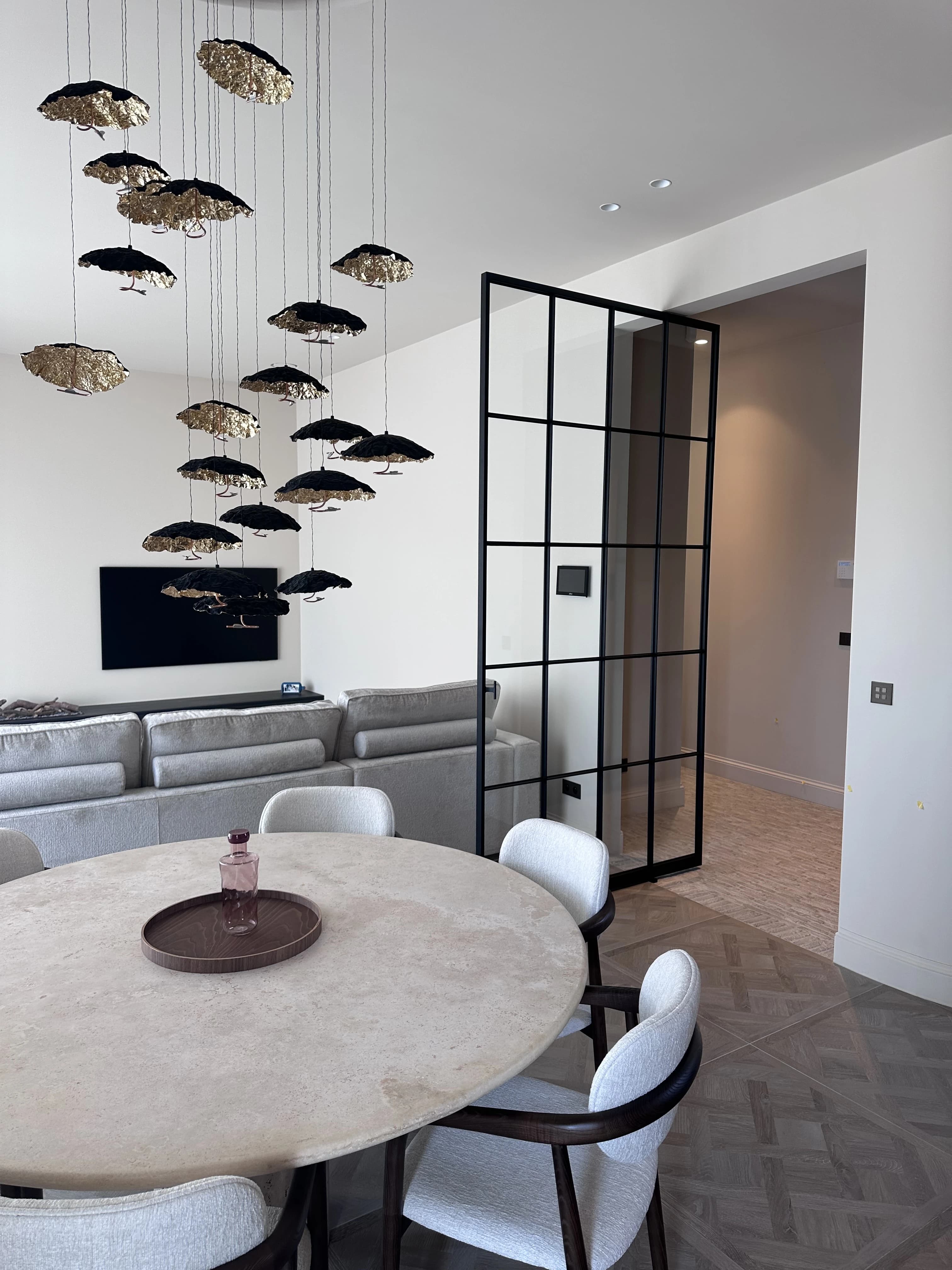 Ronde travertin eettafel met bouclé stoelen onder een sculpturale hanglamp, met zicht op de zithoek en een stalen glaswand.