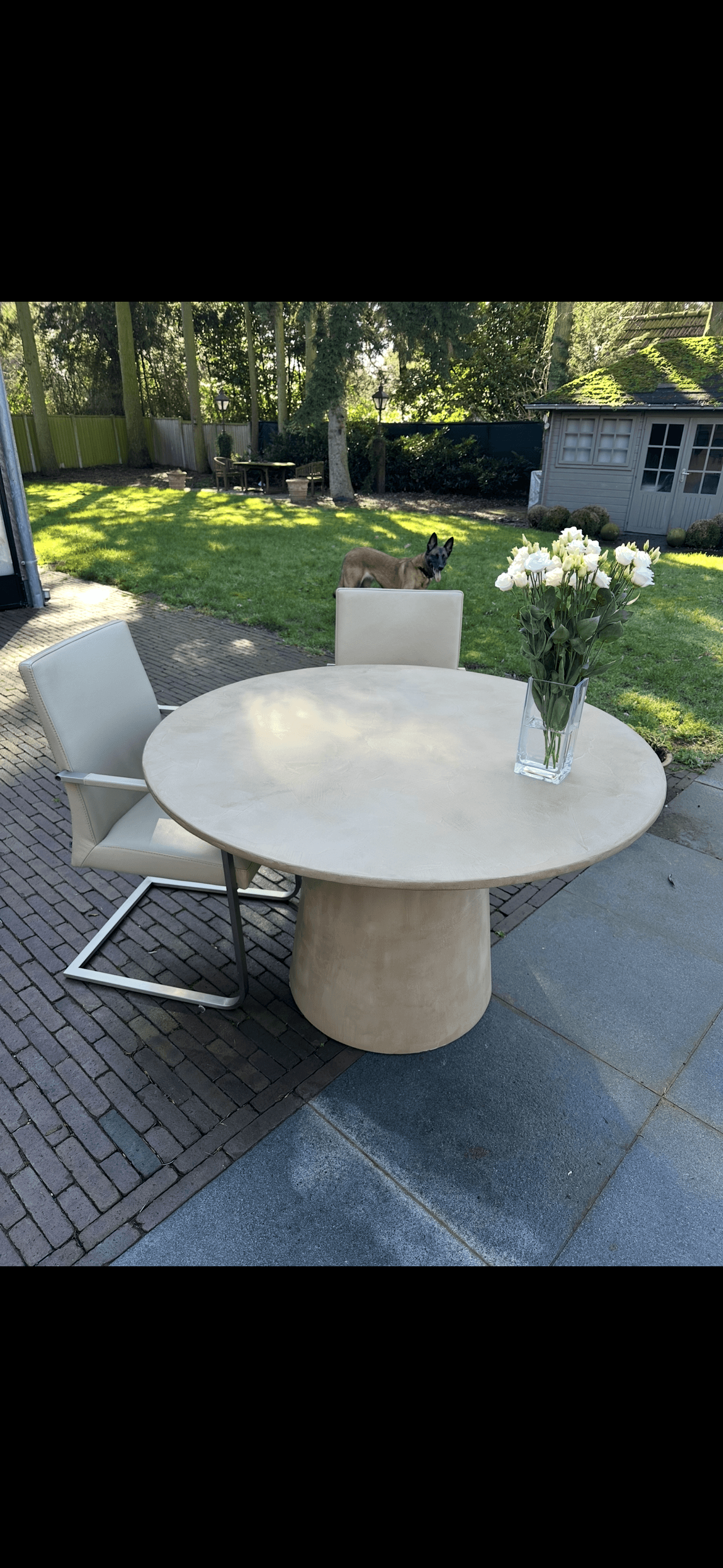 Ronde Mortex tafel met zandkleurig, naadloos blad op een zware kolomvoet, opgesteld op een terras in de tuin.